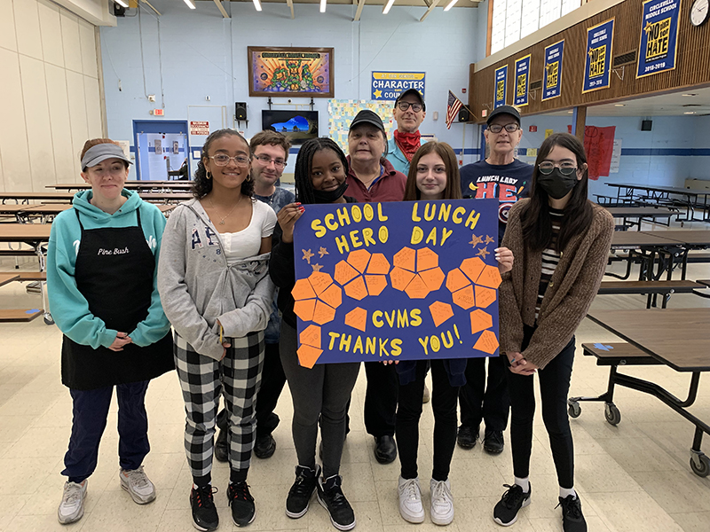 A group of five middle school students and four food service workers standing together smiling. Two of the students hold a blue and gold poster that says School Lunch Hero Day CVMS Thanks You!