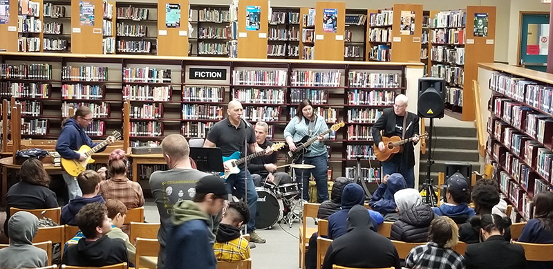 A large group of students sit with their backs facing the camera. In front of them are five adults playing musical instruments. In the background are shelves of library books.