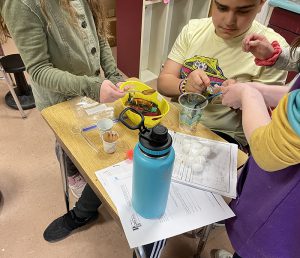 Three elementary students stand around a desk and mix oil and cocoa for an experiment.