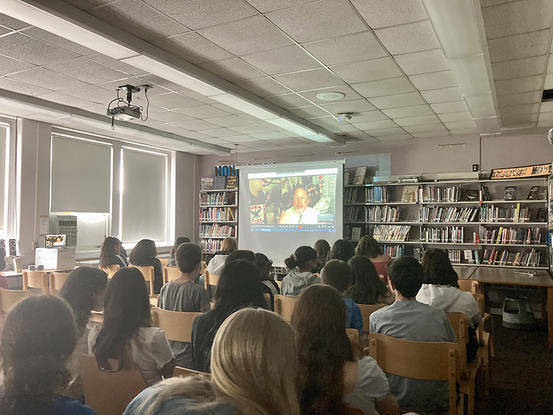 A screen at the front of the class shows a man talking. in front of the screen is a classroom filled with eighth-grade students . We see the backs of the students heads as they watch and listen to the speaker.