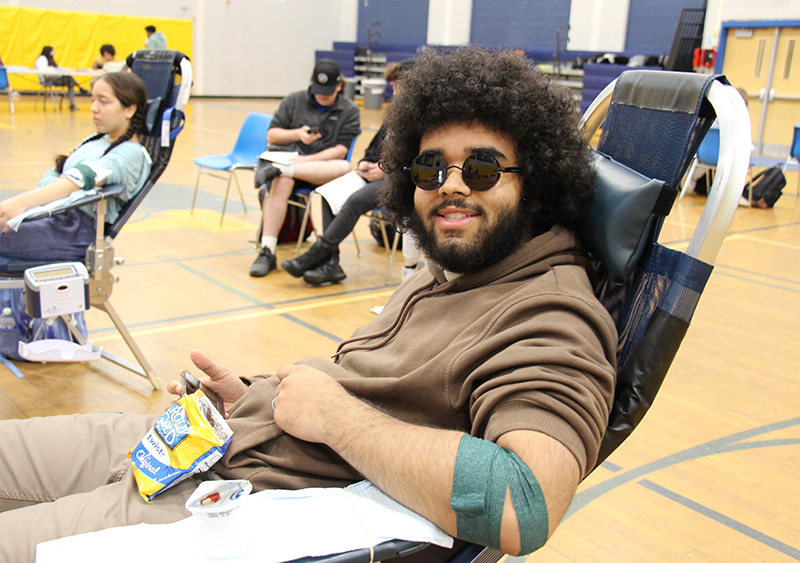 A young man with dark curly hair, beard and mustache, wearing a brown shirt and sunglasses, smiles while reclining on a cot and eating a snack.