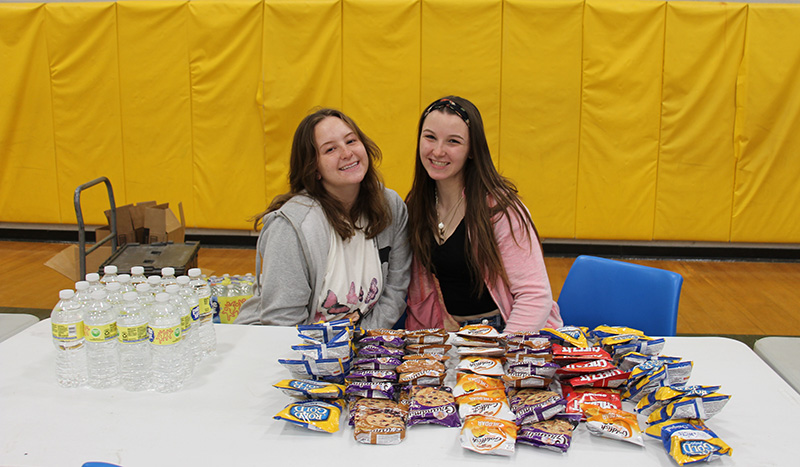 Two young women, both with long dark hair and smiling, sit behind a table that has rows of small bags of snacks on them. On the left are bottles of water all lined up.