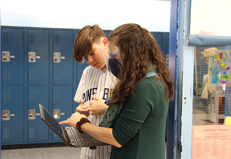 A woman in a green sweater with long dark hair and a black mask, stands in a doorway with a tall middle school student who is wearing a baseball uniform and holding a chromebook. They are both looking at the screen.