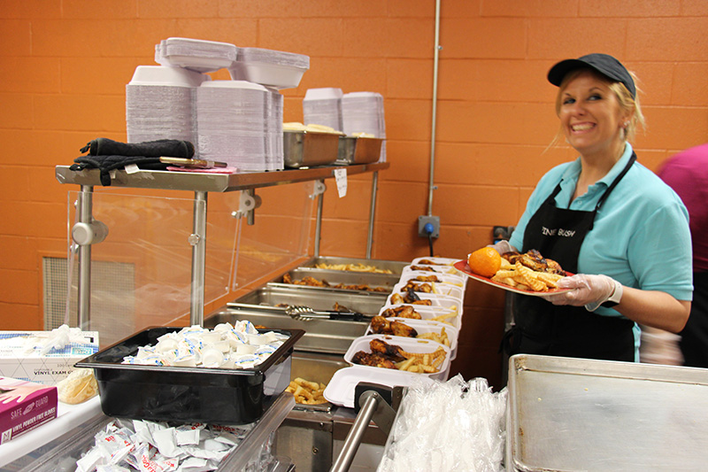 A woman in a blue shirt and black apron, wearing a black visor, holds a tray of food and smiles.