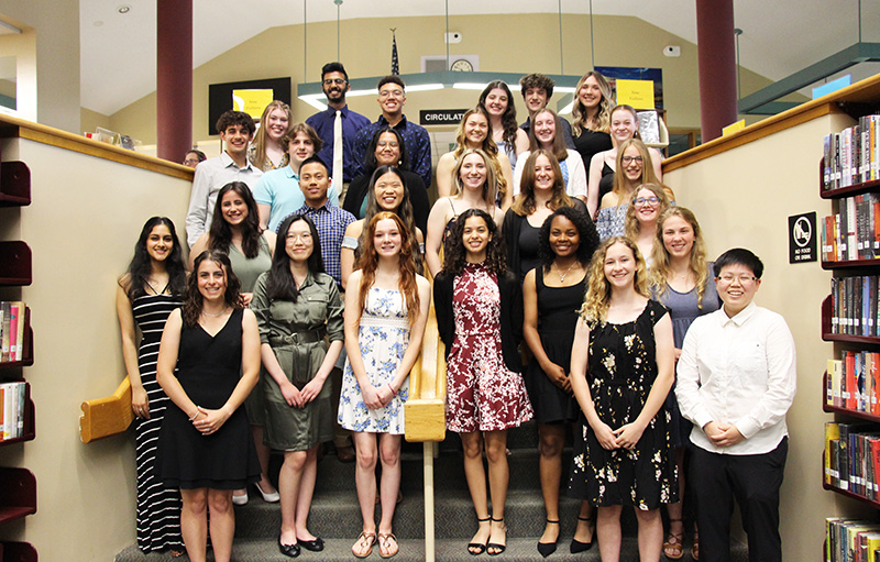 A group of 28 high school seniors stand on steps going up in a library. They are all smiling.