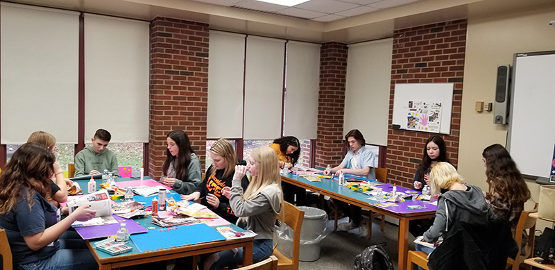 A group of high school students sit at tables that have posters and magazines on them. They are cutting things out of the magazines and gluing them onto the posters.