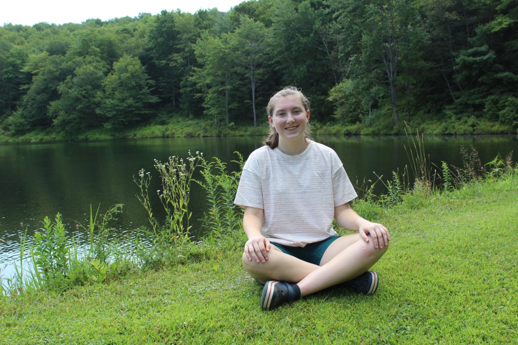 A young woman with her long hair pulled back into a ponytail sits crosslegged on the grass with a lake and trees behind her. She is wearing shorts, sneakers and a white tshirt. She is smiling.