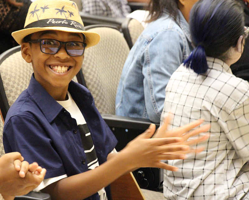 A young man wearing a navy blue shirt and a dapper straw hat claps and smiles while sitting in his seat.