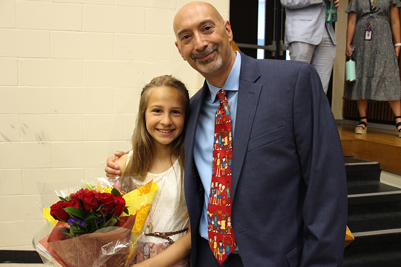 A fifth-grade student holding red flowers and wearing a white dress smiles. On the right is a man with his arm around her. He is smiling and wearing a blue suit, blue shirt and red patterned tie.