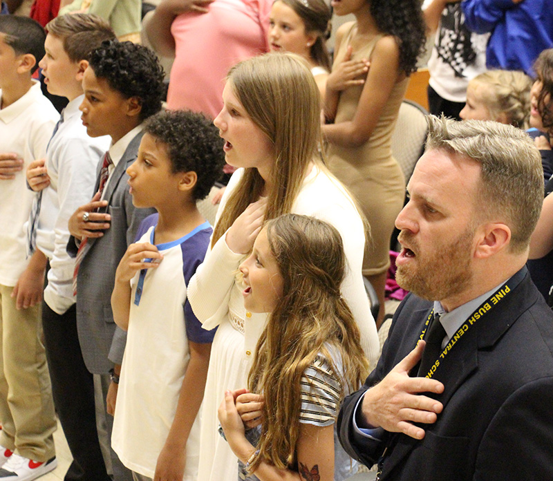 A man wearing a suit and tie stands next to a group of fifth-grade students. All are standing with their hands over their hears as they recite the Pledge of Allegience.