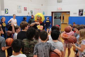 A man with a black tshirt and bright yellow fuzzy wig stands in the center of many elementary students, several have basketballs on them.