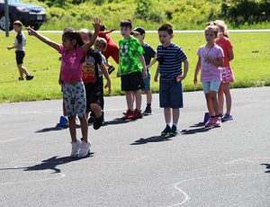 A group of elementary-age kids stand in a line. The girl at the front holds her arms up victoriously.