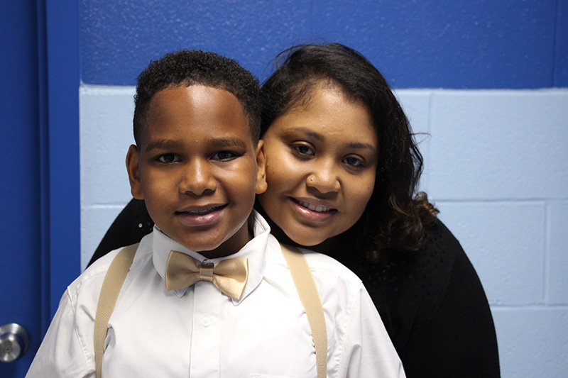 A young man in a white shirt and gold bow tie and suspenders smiles as his mom stands behind him with her head on his shoulder.