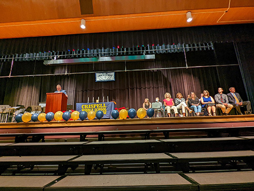 Looking up at a stage with risers in front of it. A man is standing at a wooden dais on the left. There are blue and gold balloons in front of the stage, then a group of adults and middle school students sitting to the  right.