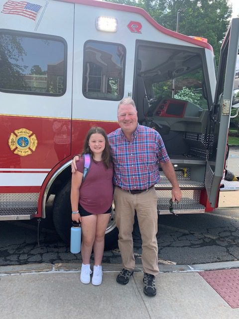 A fifth-grade girl in a reddish shirt and dark shorts and sneakers holds a water bottle. A man wearing beige pants and a blue and red plaid shirt has his arm around her shoulder. They are standing in front of a fire truck.