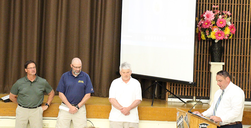 Three men stand to the left in front of a stage. A man in shirt and tie stands at a podium on the right speaking.