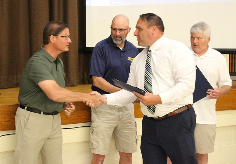 Three men stand in front of a stage. A man in a white shirt and tie shakes hands with one man and hands him a certificate as the other two look on.