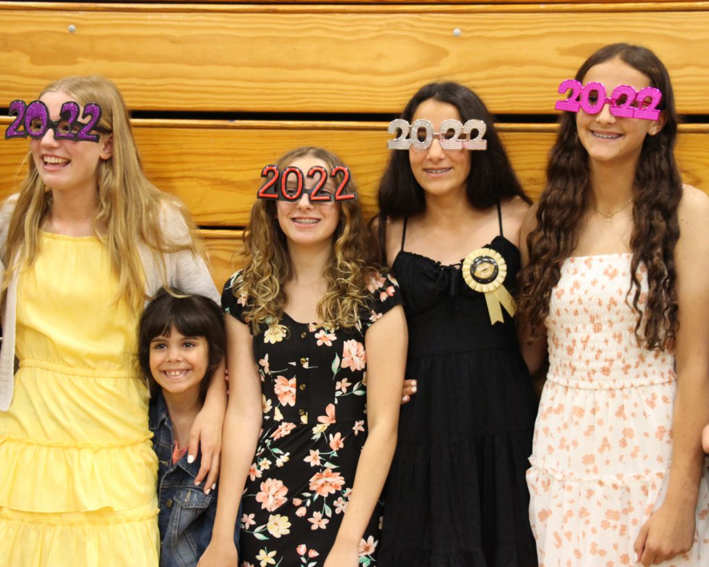 Four eighth-grade girl all wearing 2022 glasses stand in front of bleachers and smile. Next to the first and second girl on the left is a small kid poking through their arms.