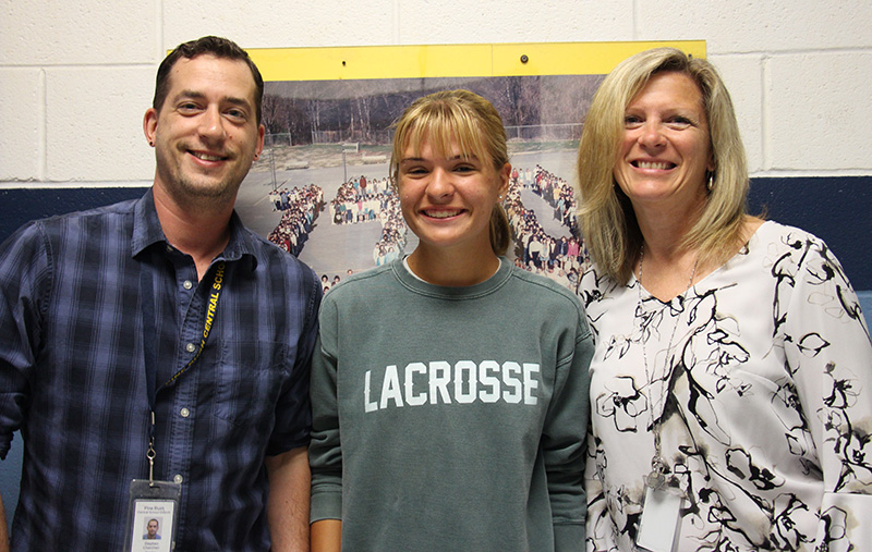 A man on the left is smiling and wearing a blue and black plaid button-down shirt. The woman on the right has medium-length blonde hair, is smiling and wearing a black and white print blouse. The young woman in the middle has her blonde hair pulled back and is smiling. She has on a green sweatshirt that says Lacrosse.