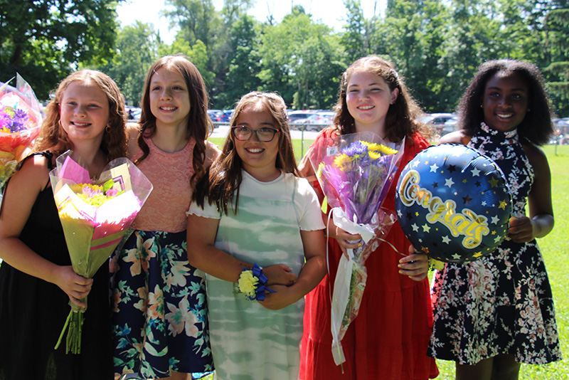 Five girls stand together, all smiling, holding balloons and flowers.