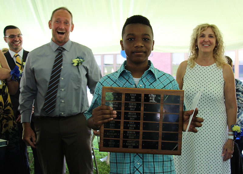 A young man holds a plaque with many different names on it. He is wearing a blue plaid shirt. Behind him are adults, two men on the left and a woman on the right.