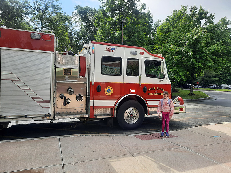 A fifth grade girl stands in front of a fire engine. She is wearing pink pants and shirt.