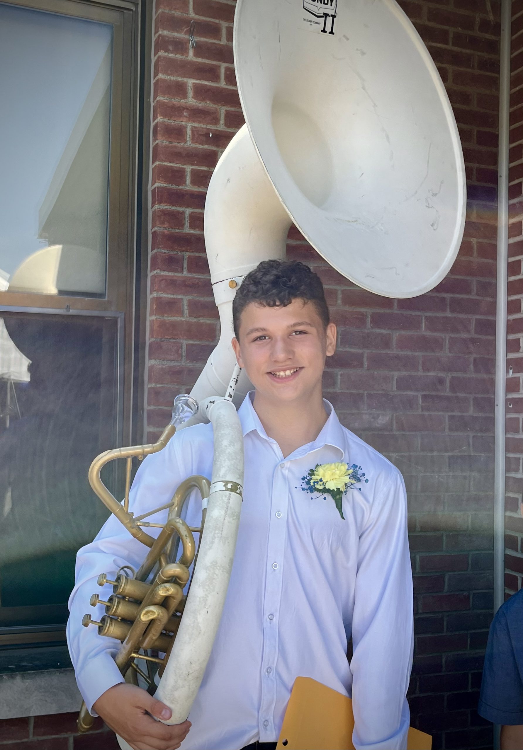 A boy wearing a white shirt and yellow boutonneire smiles in front of a building. He has a large tuba over his shoulder.