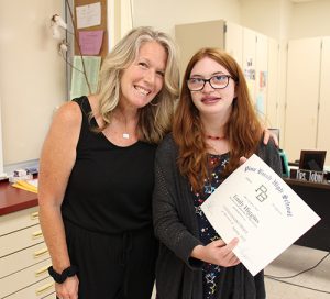 A woman with shoulder-length blonde hair dressed in a black shirt and pants has her arm around a high school girl with long reddish hair also dressed in black. She is holding a certificate and is smiling.