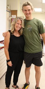 A tall high school boy wearing a green shirt smiles and stands with a woman with shoulder-length blonde hair wearing black pants and shirt.