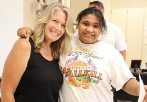 A woman with shoulder-length blonde hair dressed in a black shirt and pants has her arm around a high school girl wearing a light t-shirt. She is holding a certificate and is smiling.