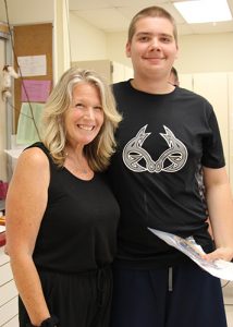 A woman with shoulder-length blonde hair dressed in a black shirt and pants has her arm around a high school boy also dressed in black. He is holding a certificate and is smiling.