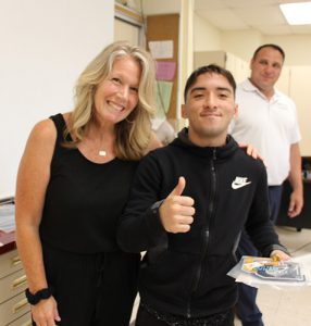 A woman with shoulder-length blonde hair dressed in a black shirt and pants has her arm around a high school boy also dressed in black. He is holding a certificate and is smiling. He also is giving a thumbs up. There is a man in the background.