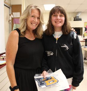 A woman with shoulder-length blonde hair dressed in a black shirt and pants has her arm around a high school girl with shoulder-length brown hair  also dressed in black. She is holding a certificate and PB Letter and is smiling.