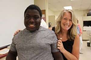 A woman with shoulder-length blonde hair dressed in a black shirt and pants has her arm around a high school boy with short dark hair, wearing a gray t-shirt. He is smiling.