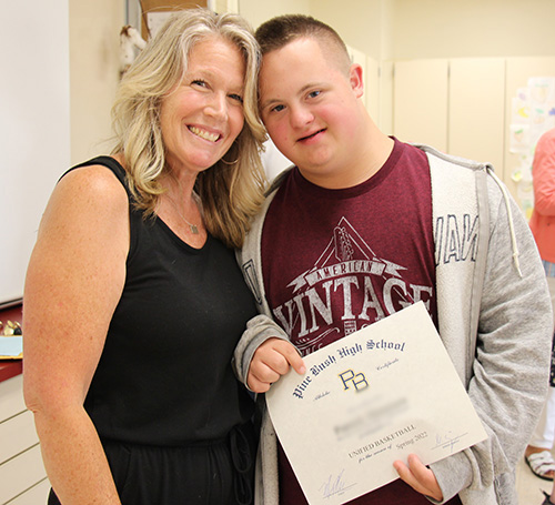 A woman with shoulder-length blonde hair dressed in a black shirt and pants has her arm around a high school boy dressed in a maroon t-shirt and gray sweatshirt. He is holding a certificate and is smiling. They have their heads leaning in together.