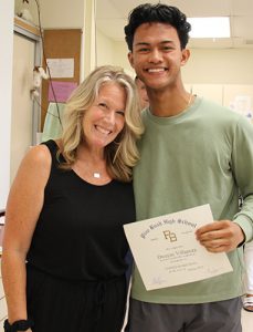 A woman with shoulder-length blonde hair dressed in a black shirt and pants has her arm around a high school boy with short dark hair and a green shirt.. He is holding a certificate and is smiling.