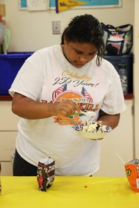 A girl with long dark hair pulled back, wearing a white tshirt with gold and blue design holds a bowl of ice cream as she puts a cherry on it.