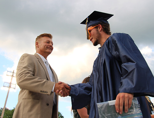 A young man in a blue cap and gown shakes the hand of a man with light colored hair and a tan suit.