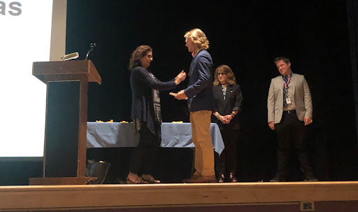 A high school senior with blonde hair, wearing a blue jacket  and tan pants, accepts a ribbon from a teacher. There are two students in the background.