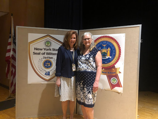 Two women stand in front of banners with shields on them. The woman on the left has long reddish hair and is wearing a navy  blue jacket and white dress. The woman on the right has a blue and white dress on and has shoulder-length light hair.