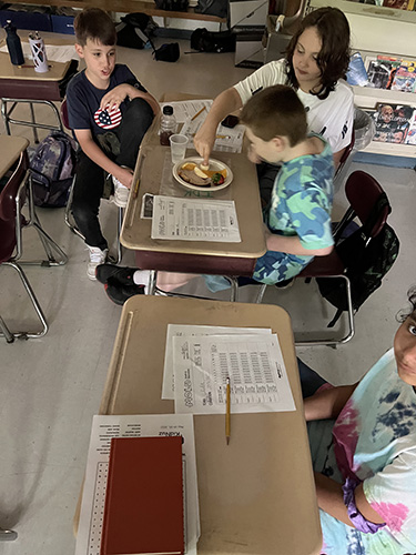 Three students sit at a desk working on a project that involves food on a plate and composting.