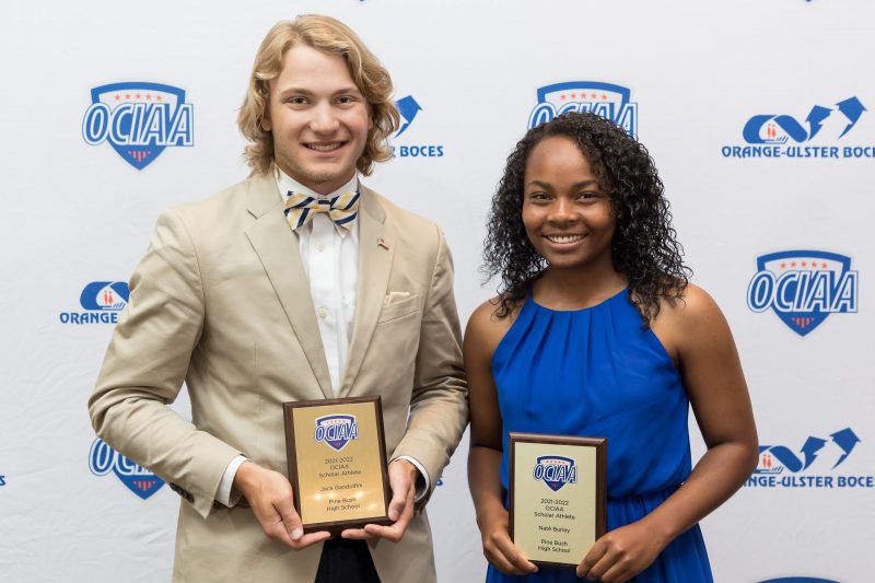 A young man with longer blonde hair, wearing a white shirt, beige jacket and beige and black bow tie stands on the right. On the left is a young woman with longer black hair, wearing a royal blue dress. Both are smiling. They are each holding gold plaques for being named the senior scholar-athletes from Pine Bush High School, given by the Orange County Interscholastic Athletic Association.