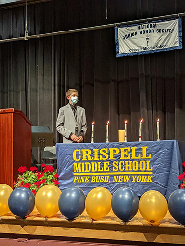 A young man wearing a jacket and a blue mask stands behind a table that contains five lit candles on it. There is a blue table cloth that says Crispell Middle School Pine Bush, New York.