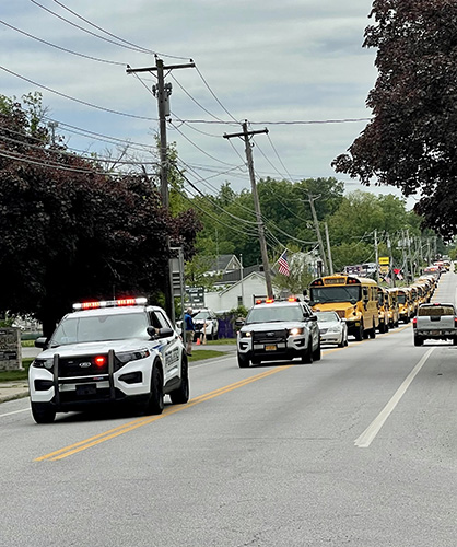 A line of police cars with lights on and school buses come down a road.