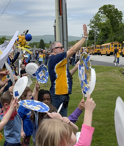 A man in a blue and gold shirt waves to the students passing on the buses. Many students are around him also waving and holding signs.