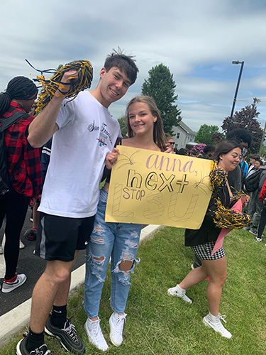 Two high scool students, a boy and girl sand outside on the grass. The boy has a blue and gold pom pom and the girl is holding a yellow sign that says Anna Next Stop LSU