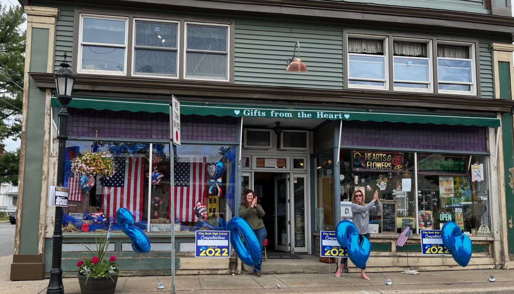 A green building is the backdrop. Two women are out in front clapping. There are large blue balloons that say 2022