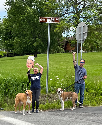 A man and woman wave. The woman is holding a large photo of a girls face on a stick. They each have a dog on a leash.