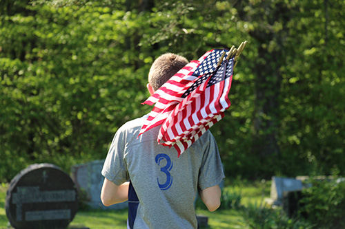 A middle school boy, wearing a gray shirt with the number 3 on it, walks away with a bunch of American flags on his shoulder. We see the back of him.