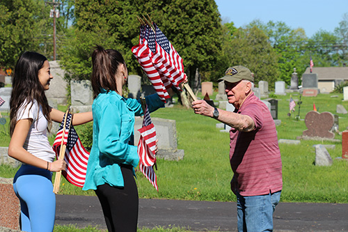 A man wearing a red shirt and a green cap has a bunch of flags in his hand and is handing them to a middle school girl wearing black pants and a green shirt. Another girl with long dark hair, light shirt and light blue pants is standing with them